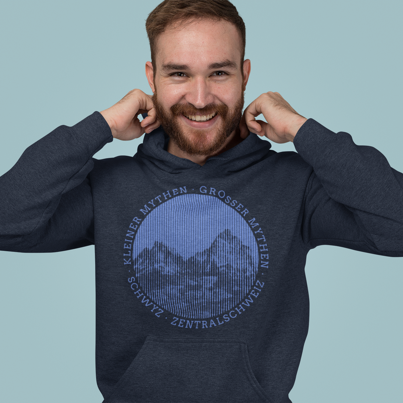 Ein junger, lachender Mann mit Bart in einem grau melierten Hoodie. Der Hoodie ist mit einem Berg bedruckt.